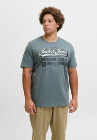 Siva majica s okruglim izrezom, kratkim rukavima i bijelim grafičkim tekstom "Jack & Jones Superior Quality" na prednjoj strani. U kombinaciji sa svijetlosmešavim hlačama.