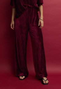 Burgundský dvoudílný outfit obsahující kalhoty s širokými nohavicemi a volný top s vertikálními texturálními pruhy, doplněný černými žabkami na světlé pleti.