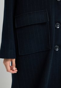 Dunkelblauer Wollmantel mit vertikalem Streifenmuster, featuring einer großen aufgesetzten Tasche und Knöpfen entlang der Vorderseite, die eine glatte Textur zeigt.