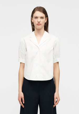 Femme portant un chemisier blanc à manches courtes avec un imprimé floral bleu clair et un pantalon large noir, debout devant un fond uni.