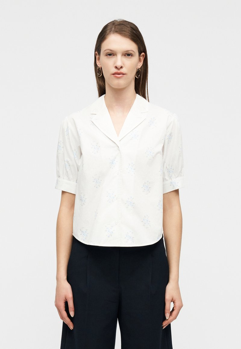 Γυναίκα που φοράει λευκό πουκάμισο με κοντά μανίκια και κουμπιά, με ανοιχτό μπλε λουλουδάτο σχέδιο, και μαύρο παντελόνι με φαρδιά πόδια, στέκεται μπροστά σε απλό φόντο.