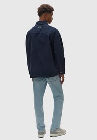 Giacca blu navy con una vestibilità rilassata, polsini con bottoni e dettaglio etichetta sulla schiena, abbinata a jeans blu chiaro e sneakers di tonalità neutra.