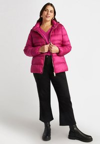 Giacca a vento magenta con chiusura zip e cappuccio, caratterizzata da una texture trapuntata e tasche laterali. Indossata sopra un top rosa abbinato e pantaloni neri.