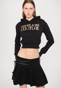 Svart kort hoodie med guld "Versace Jeans Couture" logotyp, matchad med en svart plisserad kjol med dubbla läderremmar och spännen.