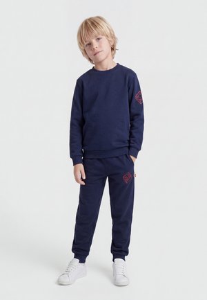 Blont barn står, bär marinblå sweatshirt och sweatpants med röda blockmönster, och vita sneakers på enfärgad vit bakgrund.