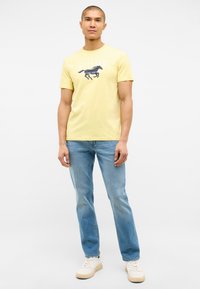 Yellow cotton t-shirt featuring a black horse graphic and white lettering, paired with light blue denim jeans and white sneakers.