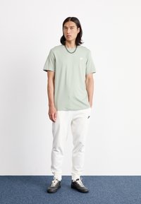 Camiseta de manga curta verde menta com um logótipo branco, combinada com calças de fato brancas e ténis pretos. Modelo de pé sobre um tapete azul.