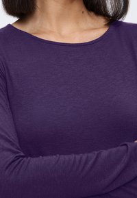 Haut à manches longues violet avec un col rond, fabriqué en tissu doux et texturé. Présente des manches ajustées et un minimum de détails de couture.