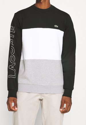 Man draagt een sweatshirt in kleurvlakken van zwart, wit en grijs met de tekst "LACOSTE" op de linkermouw en een klein krokodillenlogo op de borst.