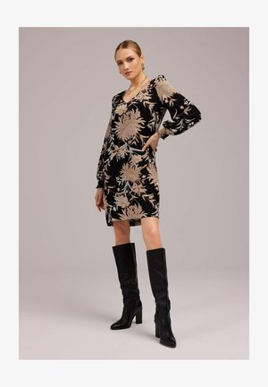 Kobieta stoi z ręką na biodrze, ubraną w czarną, kwiecistą sukienkę z długimi rękawami oraz czarne, sięgające kolan, na obcasie kozaki, na jednolitym tle.