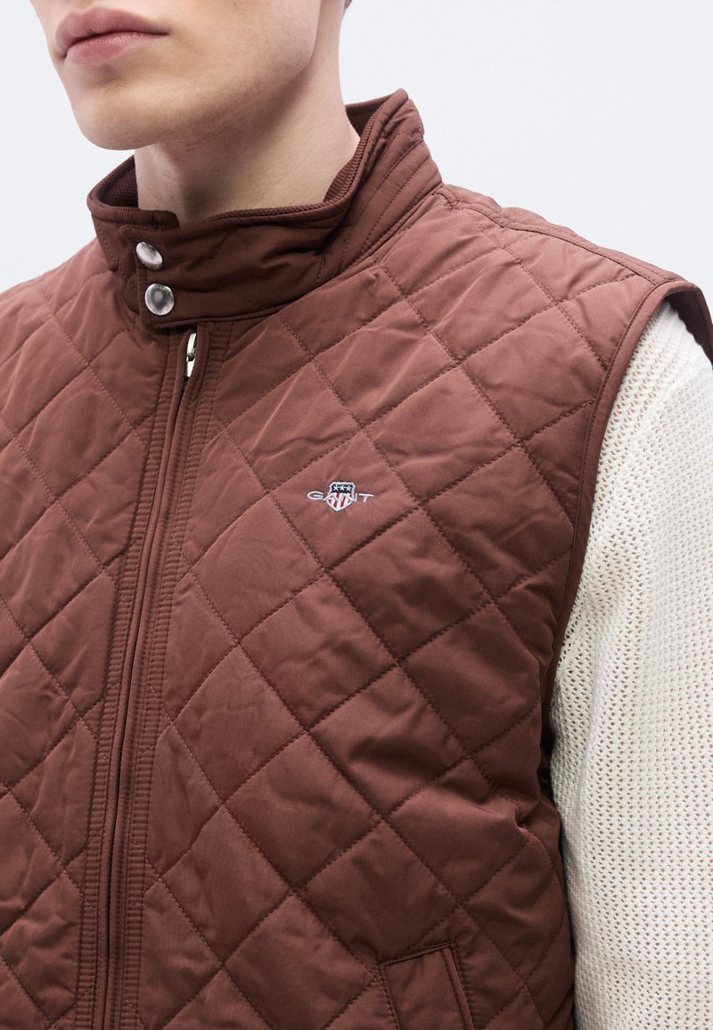 Brown quilted vest with a high collar, zip closure, and snap buttons. Features a small logo on the left chest and textured material.