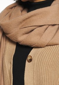 Beige ullscarf som hänger över en beige stickad cardigan med en brun knapp. Underneath en svart ribbad topp. Mjuk textur, lagerdesign.