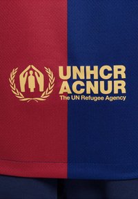Fudbalska majica s crvenim i plavim vertikalnim prugama. Sadrži zlatni logo UNHCR ACNUR i lovorov vijenac. Izrađena je od teksturiranog materijala.