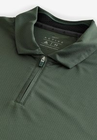 Zelena aktivna polo majica s ovratnikom na zip, teksturiranom tkaninom i mrežastim podstavom na vratu. Sadrži logo i oznaku za njegu unutar majice.