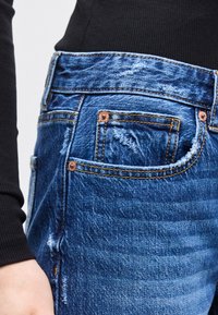 Κοντινό πλάνο μπλε τζιν με χαλκούς δυκτυωτές βίδες, κίτρινη ραφή και μικρή τσέπη, φορεμένο με μαύρο πλεκτό πάνω μέρος.