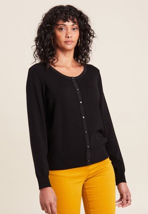 Cardigan nero con scollo rotondo, chiusura a bottoni e maniche lunghe. Presenta un bordo testurizzato e un tessuto liscio. Abbinato a pantaloni gialli.