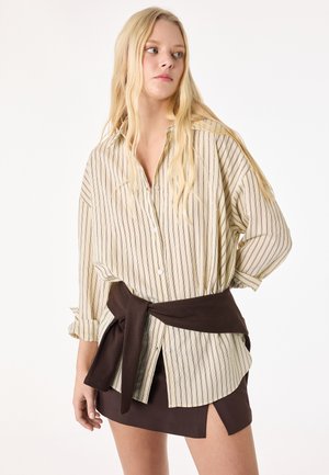 Ung kvinde med langt blondt hår iført en beige, stribet, oversized skjorte og en mørkebrun nederdel med bindebånd i taljen.