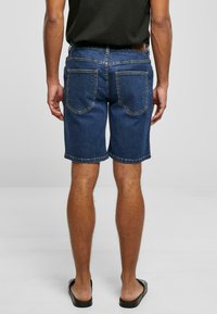 Shorts en denim bleu foncé, avec une coupe classique, jambe droite et poches arrière cousues avec un léger détail de couture. Design de ceinture plate.