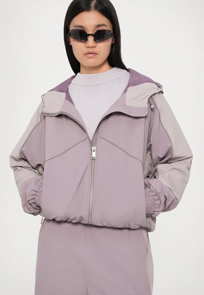 Ανοιχτό μωβ ζακέτα με φερμουάρ και κουκούλα, διαθέτει ελαστικούς μανσέτες και αντίθετο γιακά. Φοριέται πάνω από ένα υφασμάτινο ανοιχτό γκρίζο πουλόβερ.