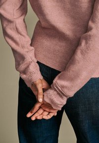 Šviesiai rožinė megzta palaidinė su tekstūrinėmis nėrinių rankovėmis, dėvima su tamsiai mėlynomis džinsinėmis kelnėmis. Rankovės pasižymi subtiliu atviru darbu.