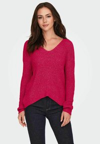 Roze gebreide trui met een V-hals, lange mouwen en een high-low zoom. Textuurpatroon voegt visuele interesse toe. Gecombineerd met donkere jeans.