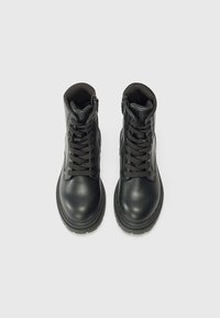 Botas de cuero negro hasta el tobillo, con punteras redondeadas, cuellos acolchados y cordones negros. Incluyen cremalleras laterales y una suela de goma texturizada.