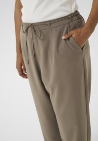 Pantalons pour adulte à taille élastique en doux tissu taupe. Dotés d'une fermeture à cordon, de poches latérales et d'une coupe ample. Texture lisse.