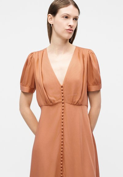 Νεαρή γυναίκα με ανοιχτόχρωμο δέρμα που φοράει φόρεμα σκουριάς με κοντά πριγκιπικά μανίκια και σειρά από κουμπιά καλυμμένα με ύφασμα μπροστά.