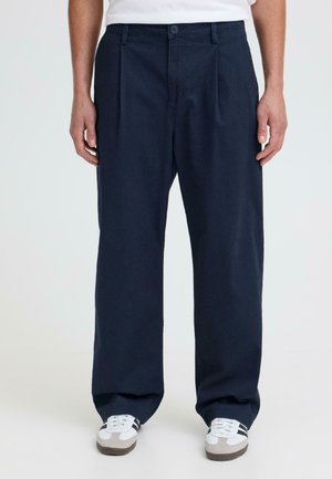 Personne portant un pantalon ample bleu marine foncé et des baskets blanches à rayures noires, debout devant un fond clair uni.