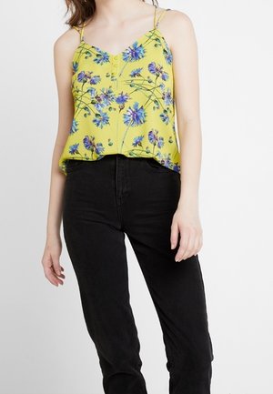 Kobieta w żółtym kwiatowym topie na cienkich ramiączkach w połączeniu z czarnymi spodniami z wysokim stanem na białym tle.