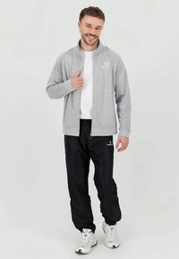 Lichtgrijze zip-up jas van textuurstof, gecombineerd met zwarte sweatpants. Witte shirt eronder en witte sport schoenen.