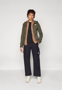 Maslinasto zelena jakna za vanjsku upotrebu preko smeđe unutarnje jakne, uparena s crnim cargo hlačama i svijetlim tenisicama.