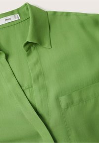 Chemise verte à col avec un décolleté en V et une poche poitrine, en tissu lisse, montrant des coutures détaillées près du col et de la poche.