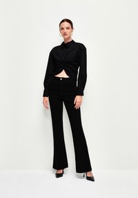 Sort kortærmet knap-op skjorte med en snoet front, parret med sorte flare jeans. Modellen har sorte højhælede sko og hoop øreringe på.