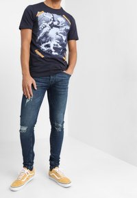 Camiseta gráfica azul marino con un diseño claro, combinada con jeans ajustados y desgastados. El atuendo se completa con zapatillas amarillas.
