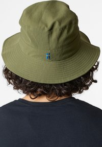 Cappello a secchiello verde oliva in cotone, con una larga visiera e un dettaglio della bandiera blue e gialla, mostrato da dietro su una persona che indossa una shirt nera.