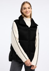 Czarna puchowa kamizelka z wysokim kołnierzem i przednim zamkiem. Zawiera kontrastowe szycie i napy. Noszona na białym swetrze z prążkowanej tkaniny.