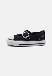 Zapato de lona negro con una puntera de goma blanca, detalle de hebilla plateada y costuras blancas. Presenta un diseño de perfil bajo y rayas en la suela de color negro.
