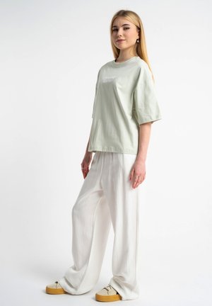 Młoda kobieta o długich blond włosach, ubrana w luźny jasnooliwkowy T-shirt, szerokie białe spodnie i beżowe trampki na białym tle.