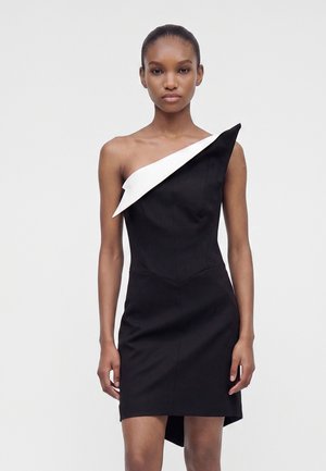 Femme portant une robe noire sans manches avec un détail blanc asymétrique rabattable sur une épaule, sur un fond clair uni.