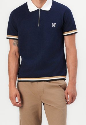 Marineblå kortærmet polo med hvid krave, halv lynlås, guld- og hvide striber på ærmer og kant, båret med beige bukser.