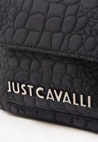 Sac à main noir matelassé avec un motif texturé, présentant le nom de marque en relief "JUST CAVALLI" en lettres métalliques sur le devant.