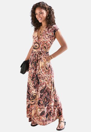 Frau lächelnd, trägt ein buntes Maxikleid mit Paisley-Muster und kurzen Ärmeln, hält eine schwarze Clutch und trägt schwarze Sandalen.