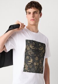 Weißes Baumwoll-T-Shirt mit einem großen schwarzen und goldenen Blumendesign in der Mitte. Das Modell trägt eine schwarze Tasche über der Schulter.