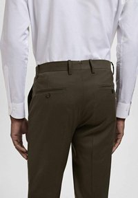 Achteraanzicht van een persoon in een donkerbruin broek met knoopzakken en een wit overhemd met lange mouwen.
