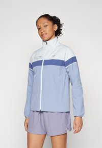 Giacca zip-up azzurro chiaro con colletto bianco e striscia blu navy sul petto. Presenta maniche lunghe e un design sportivo. Indossata con pantaloni corti viola.