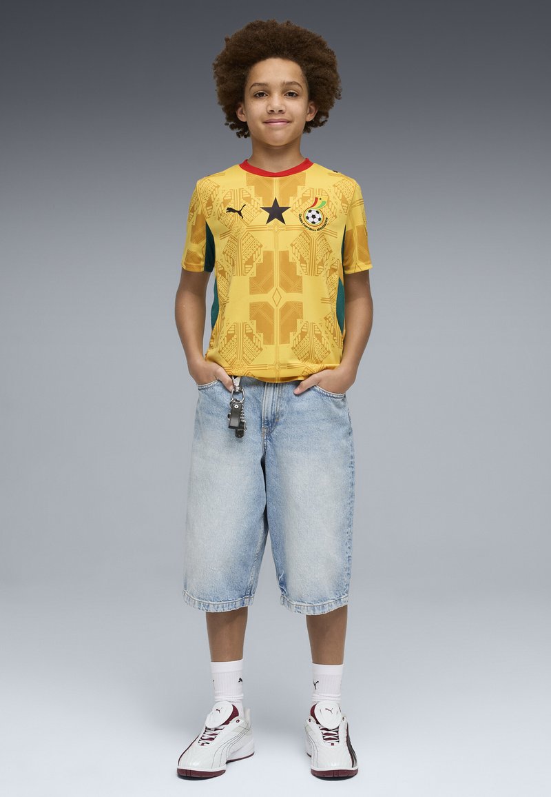 Enfant avec coiffure afro portant un maillot de football jaune du Ghana, un short en denim clair, des chaussettes blanches Puma et des baskets blanches avec des détails bordeaux.