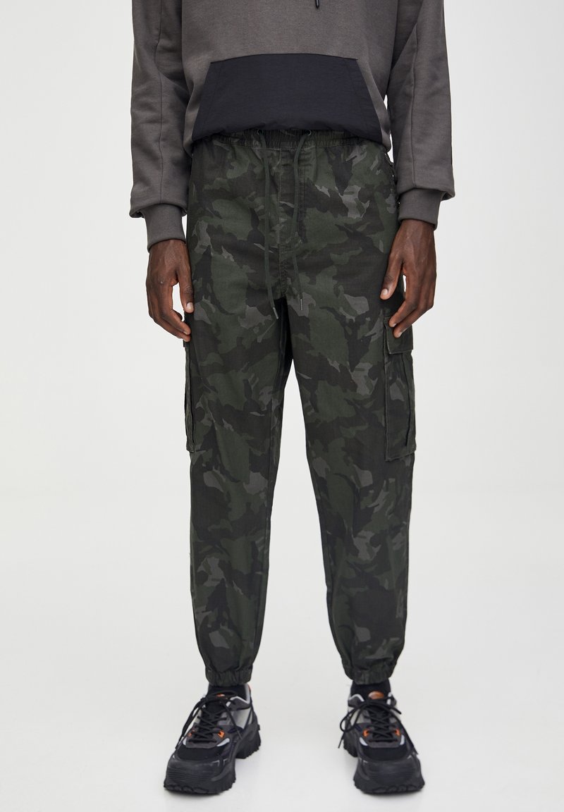 Homme portant un pantalon cargo camouflage vert foncé et noir, un sweat à capuche gris, et des baskets noires à semelles épaisses.
