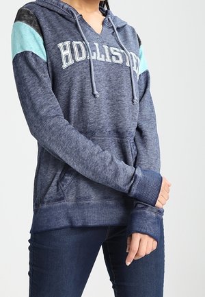 Persona vistiendo una sudadera con capucha azul de Hollister con detalles en parche azul verdoso y oscuro en las mangas, combinada con jeans azul oscuro, con los brazos cruzados a la altura de la cintura.