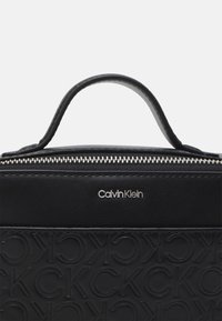 Borsa in pelle nera con logo impresso, cerniera in argento e manico circolare. Presenta un design testurizzato con lettere ripetute.